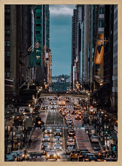 A high-angle, vertical photograph looking down a bustling New York City street at dusk, framed by towering skyscrapers. The street below is packed with cars, their headlights creating a warm golden glow, contrasting with the cool, dark blue tones of the twilight sky and buildings. Poster
