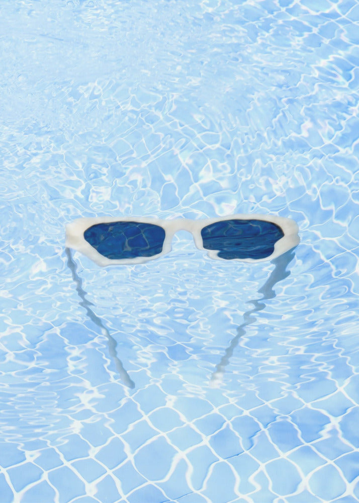 A pair of white-framed sunglasses with dark lenses floats on the surface of a bright blue swimming pool. The rippling water distorts the view of the tiled pool floor, and the sunglasses cast a faint shadow underneath. Print