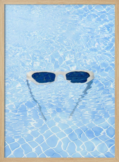 A pair of white-framed sunglasses with dark lenses floats on the surface of a bright blue swimming pool. The rippling water distorts the view of the tiled pool floor, and the sunglasses cast a faint shadow underneath. Print