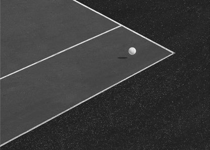 A minimalist black and white overhead photograph of a white ball resting in the corner of a dark tennis or paddle court. The composition focuses on the strong geometric angles created by the white boundary lines against the dark, textured asphalt, all within a silver frame. Artwork