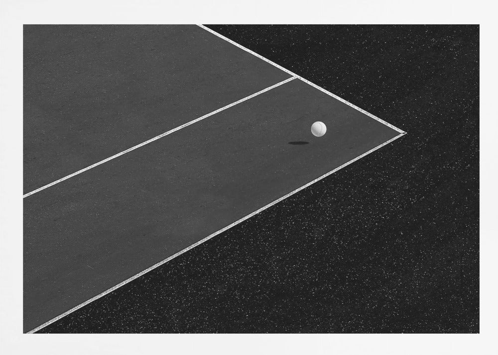 A minimalist black and white overhead photograph of a white ball resting in the corner of a dark tennis or paddle court. The composition focuses on the strong geometric angles created by the white boundary lines against the dark, textured asphalt, all within a silver frame. Artwork