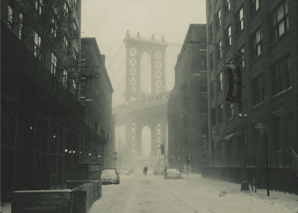 A framed, vintage-style black and white photograph capturing a snowy day in DUMBO, Brooklyn. The view looks down a snow-covered street lined with dark buildings, perfectly framing the Manhattan Bridge in the foggy distance. A few cars are parked along the street, and snow is visibly falling, creating a quiet, atmospheric winter scene. Decor