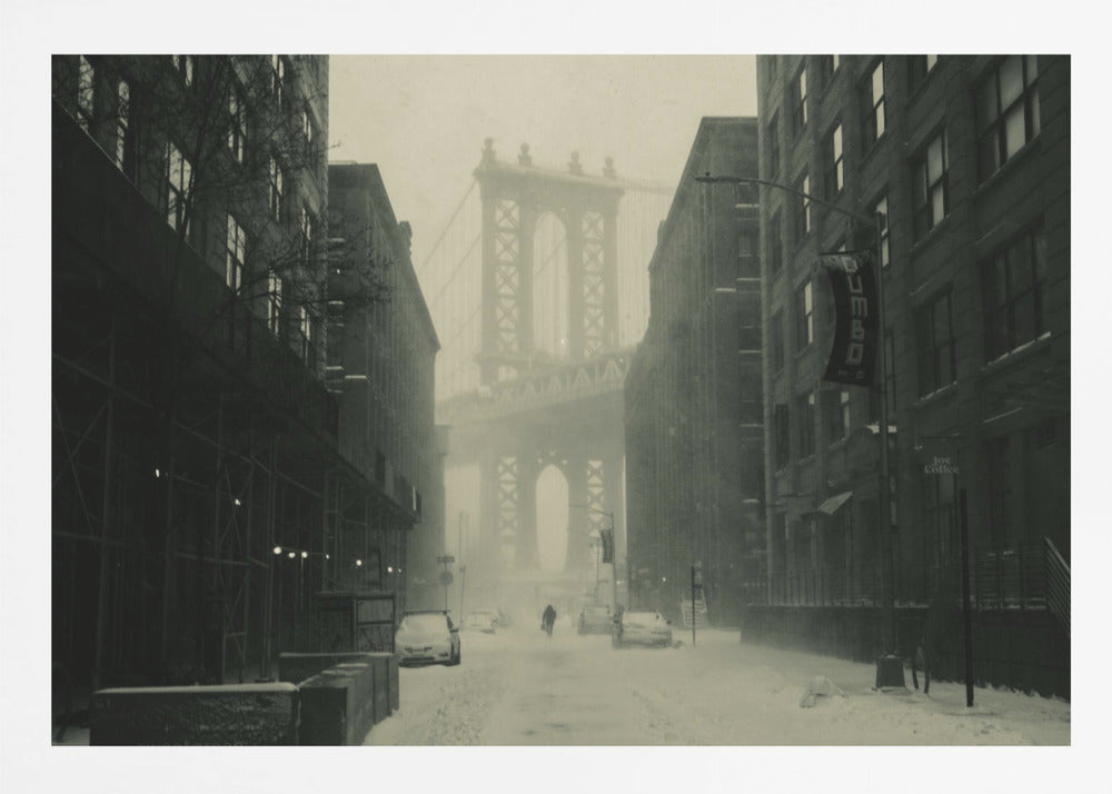 A framed, vintage-style black and white photograph capturing a snowy day in DUMBO, Brooklyn. The view looks down a snow-covered street lined with dark buildings, perfectly framing the Manhattan Bridge in the foggy distance. A few cars are parked along the street, and snow is visibly falling, creating a quiet, atmospheric winter scene. Decor
