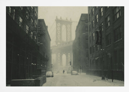 A framed, vintage-style black and white photograph capturing a snowy day in DUMBO, Brooklyn. The view looks down a snow-covered street lined with dark buildings, perfectly framing the Manhattan Bridge in the foggy distance. A few cars are parked along the street, and snow is visibly falling, creating a quiet, atmospheric winter scene. Decor
