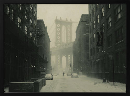 A framed, vintage-style black and white photograph capturing a snowy day in DUMBO, Brooklyn. The view looks down a snow-covered street lined with dark buildings, perfectly framing the Manhattan Bridge in the foggy distance. A few cars are parked along the street, and snow is visibly falling, creating a quiet, atmospheric winter scene. Decor