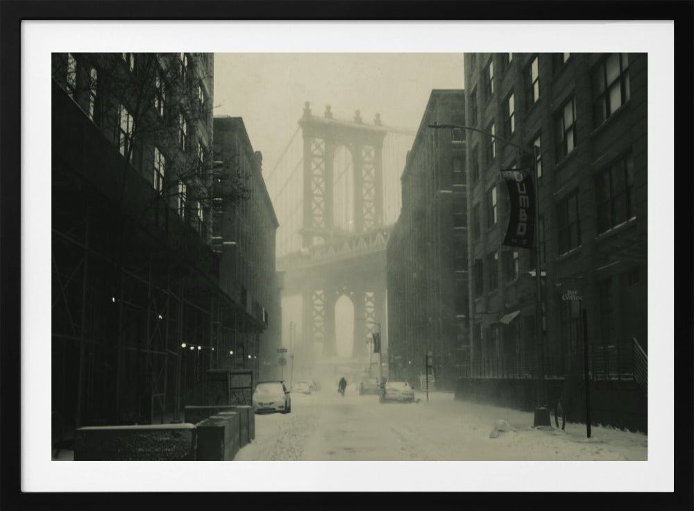 A framed, vintage-style black and white photograph capturing a snowy day in DUMBO, Brooklyn. The view looks down a snow-covered street lined with dark buildings, perfectly framing the Manhattan Bridge in the foggy distance. A few cars are parked along the street, and snow is visibly falling, creating a quiet, atmospheric winter scene. Decor