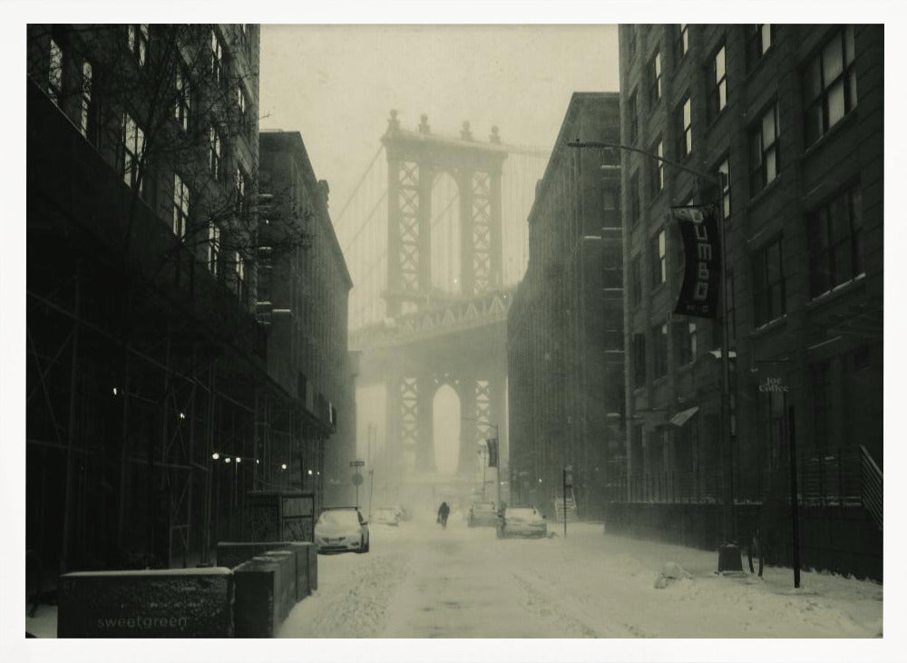 A framed, vintage-style black and white photograph capturing a snowy day in DUMBO, Brooklyn. The view looks down a snow-covered street lined with dark buildings, perfectly framing the Manhattan Bridge in the foggy distance. A few cars are parked along the street, and snow is visibly falling, creating a quiet, atmospheric winter scene. Decor