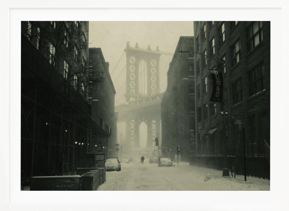 A framed, vintage-style black and white photograph capturing a snowy day in DUMBO, Brooklyn. The view looks down a snow-covered street lined with dark buildings, perfectly framing the Manhattan Bridge in the foggy distance. A few cars are parked along the street, and snow is visibly falling, creating a quiet, atmospheric winter scene. Decor