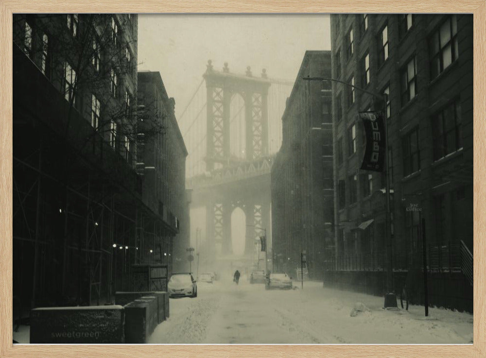 A framed, vintage-style black and white photograph capturing a snowy day in DUMBO, Brooklyn. The view looks down a snow-covered street lined with dark buildings, perfectly framing the Manhattan Bridge in the foggy distance. A few cars are parked along the street, and snow is visibly falling, creating a quiet, atmospheric winter scene. Decor