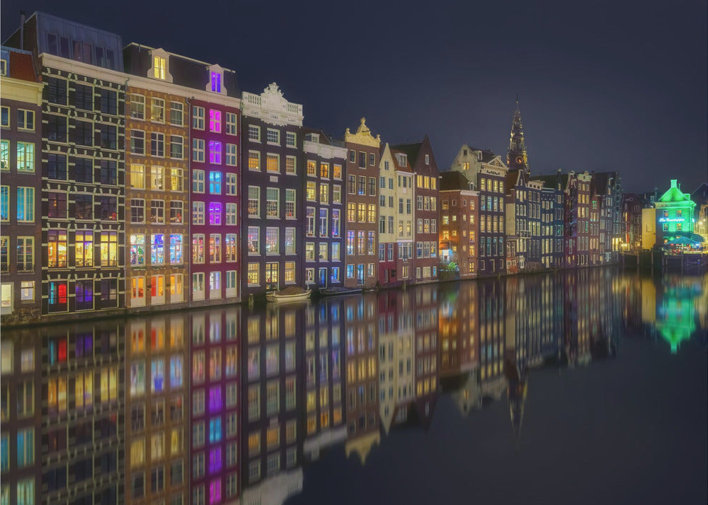 A vibrant nighttime photograph of colorful canal houses in Amsterdam, with their glowing lights beautifully reflected in the still water of the canal below, all framed in silver. Print