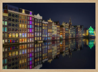 A vibrant nighttime photograph of colorful canal houses in Amsterdam, with their glowing lights beautifully reflected in the still water of the canal below, all framed in silver. Print