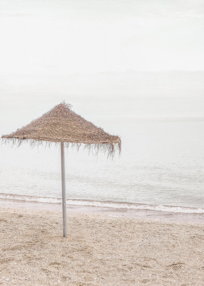 A minimalist photograph of a solitary thatched umbrella on a sandy beach, with calm, grey water and a hazy, overcast sky in the background, creating a serene and tranquil coastal scene. Poster