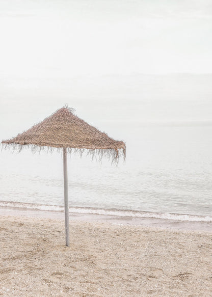 A minimalist photograph of a solitary thatched umbrella on a sandy beach, with calm, grey water and a hazy, overcast sky in the background, creating a serene and tranquil coastal scene. Poster