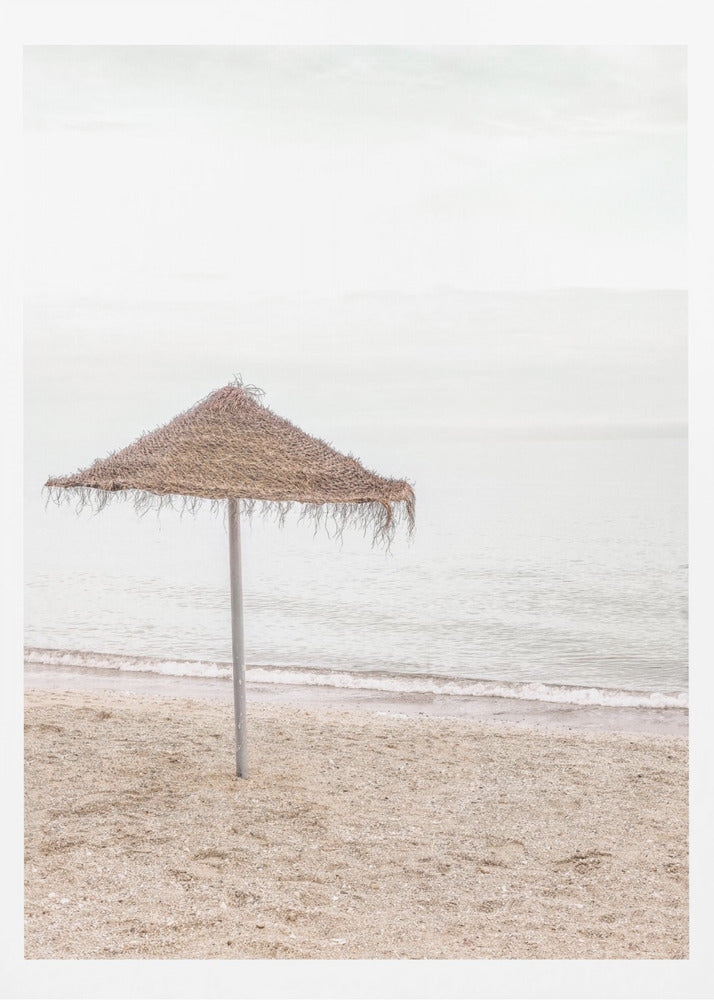 A minimalist photograph of a solitary thatched umbrella on a sandy beach, with calm, grey water and a hazy, overcast sky in the background, creating a serene and tranquil coastal scene. Poster