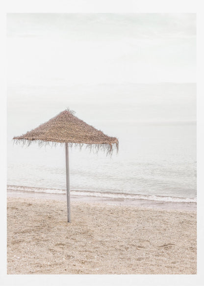 A minimalist photograph of a solitary thatched umbrella on a sandy beach, with calm, grey water and a hazy, overcast sky in the background, creating a serene and tranquil coastal scene. Poster