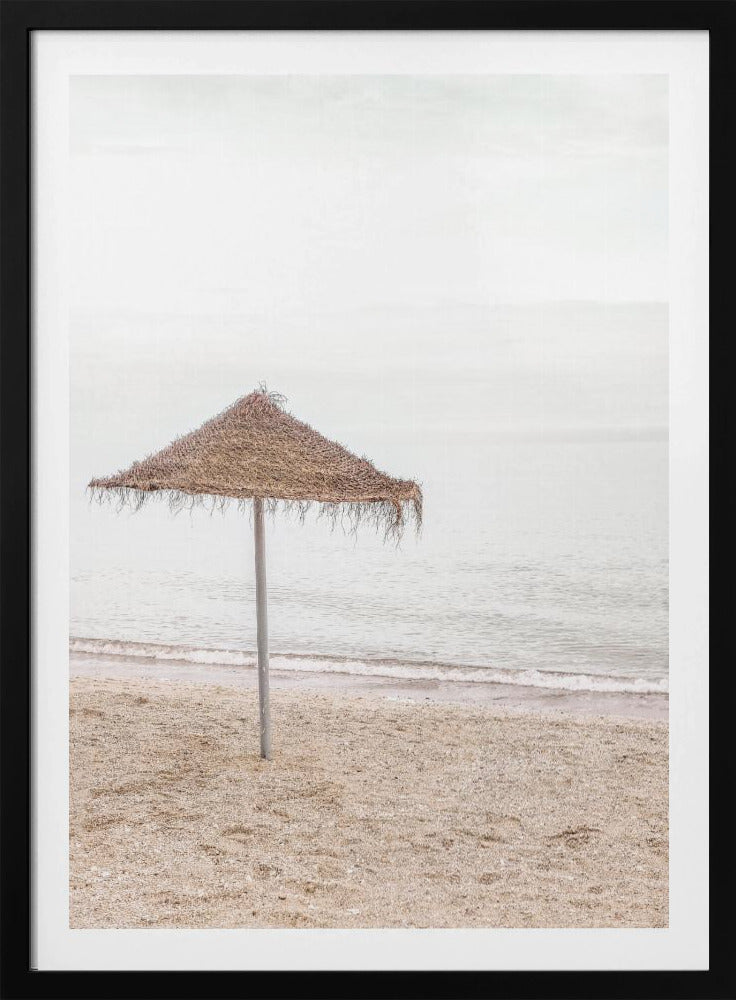 A minimalist photograph of a solitary thatched umbrella on a sandy beach, with calm, grey water and a hazy, overcast sky in the background, creating a serene and tranquil coastal scene. Poster