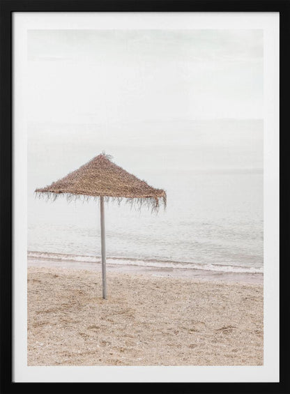 A minimalist photograph of a solitary thatched umbrella on a sandy beach, with calm, grey water and a hazy, overcast sky in the background, creating a serene and tranquil coastal scene. Poster