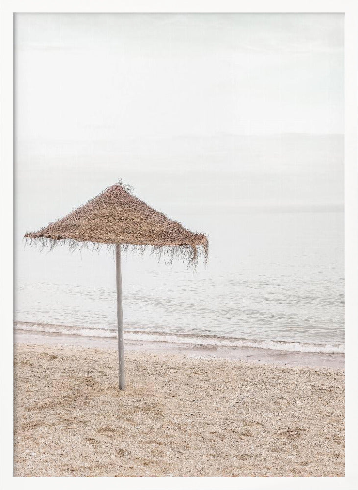 A minimalist photograph of a solitary thatched umbrella on a sandy beach, with calm, grey water and a hazy, overcast sky in the background, creating a serene and tranquil coastal scene. Poster