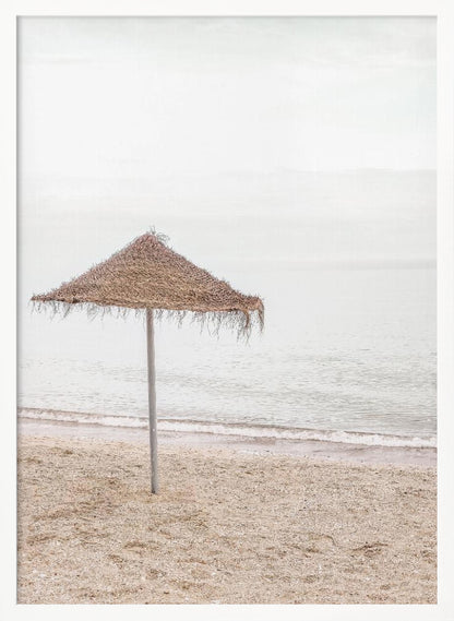 A minimalist photograph of a solitary thatched umbrella on a sandy beach, with calm, grey water and a hazy, overcast sky in the background, creating a serene and tranquil coastal scene. Poster