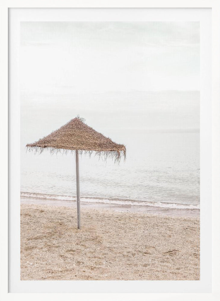A minimalist photograph of a solitary thatched umbrella on a sandy beach, with calm, grey water and a hazy, overcast sky in the background, creating a serene and tranquil coastal scene. Poster
