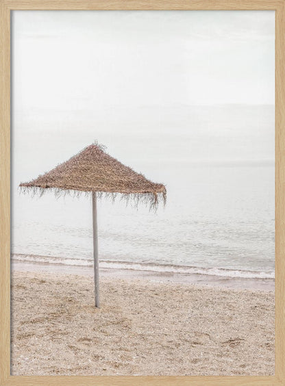 A minimalist photograph of a solitary thatched umbrella on a sandy beach, with calm, grey water and a hazy, overcast sky in the background, creating a serene and tranquil coastal scene. Poster