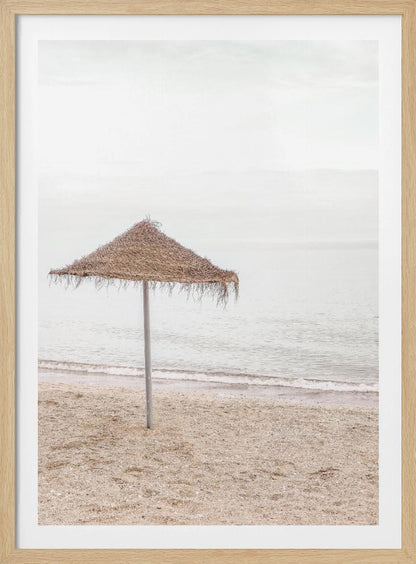 A minimalist photograph of a solitary thatched umbrella on a sandy beach, with calm, grey water and a hazy, overcast sky in the background, creating a serene and tranquil coastal scene. Poster