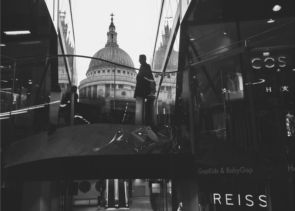 A framed, black and white photograph capturing the reflection of St. Paul's Cathedral in the glass of a modern shopping center. A person's silhouette stands on an interior balcony, juxtaposed against the historic dome, while the mall's interior and store signs are visible in the foreground. Print