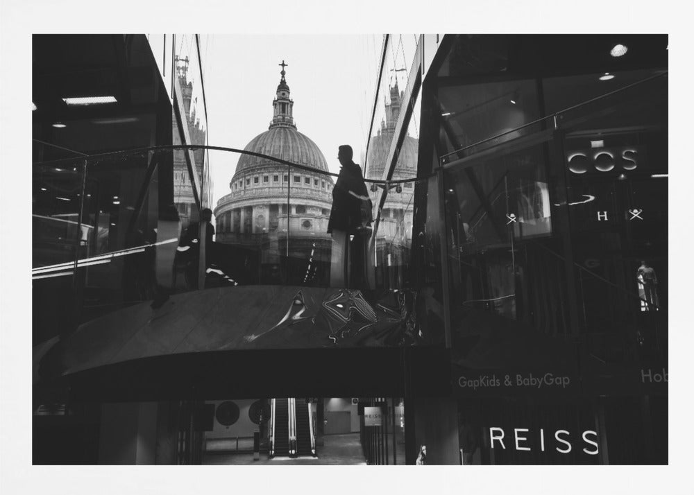 A framed, black and white photograph capturing the reflection of St. Paul's Cathedral in the glass of a modern shopping center. A person's silhouette stands on an interior balcony, juxtaposed against the historic dome, while the mall's interior and store signs are visible in the foreground. Print