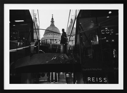 A framed, black and white photograph capturing the reflection of St. Paul's Cathedral in the glass of a modern shopping center. A person's silhouette stands on an interior balcony, juxtaposed against the historic dome, while the mall's interior and store signs are visible in the foreground. Print