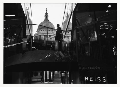 A framed, black and white photograph capturing the reflection of St. Paul's Cathedral in the glass of a modern shopping center. A person's silhouette stands on an interior balcony, juxtaposed against the historic dome, while the mall's interior and store signs are visible in the foreground. Print