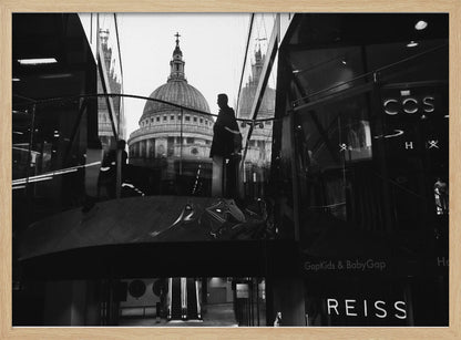 A framed, black and white photograph capturing the reflection of St. Paul's Cathedral in the glass of a modern shopping center. A person's silhouette stands on an interior balcony, juxtaposed against the historic dome, while the mall's interior and store signs are visible in the foreground. Print
