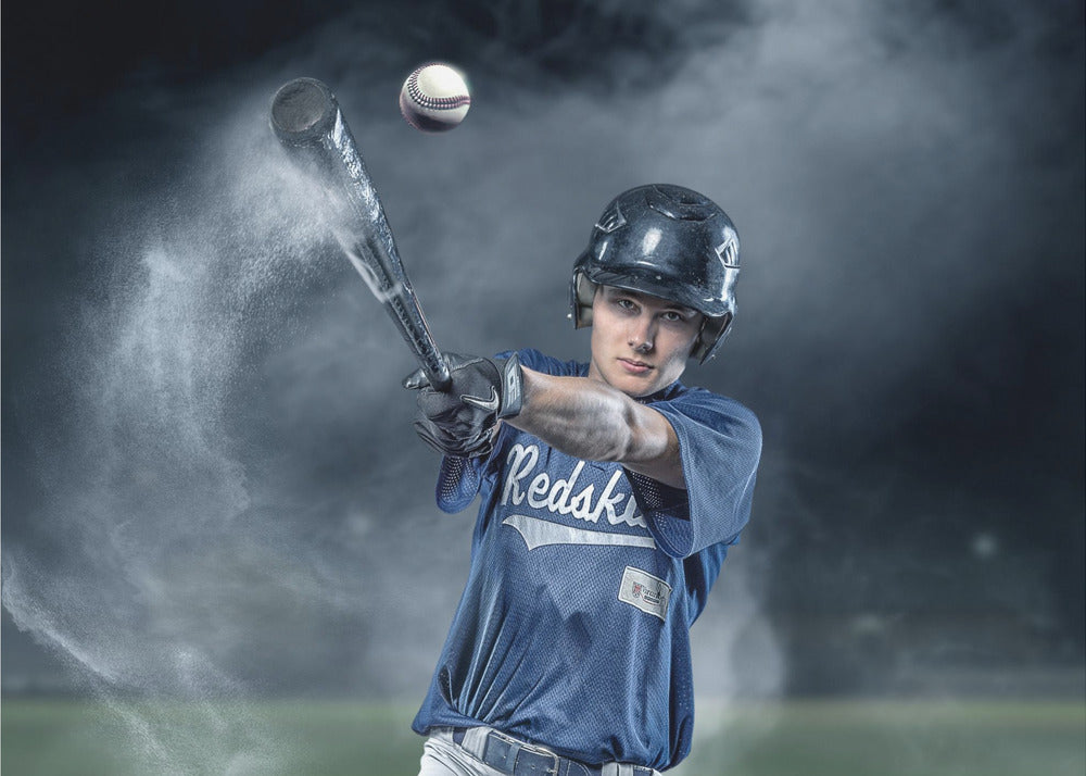 A dramatic, framed portrait of a young baseball player in a blue jersey and helmet, captured in the moment of hitting a baseball, with a cloud of dust exploding from the bat against a dark, smoky background. Poster