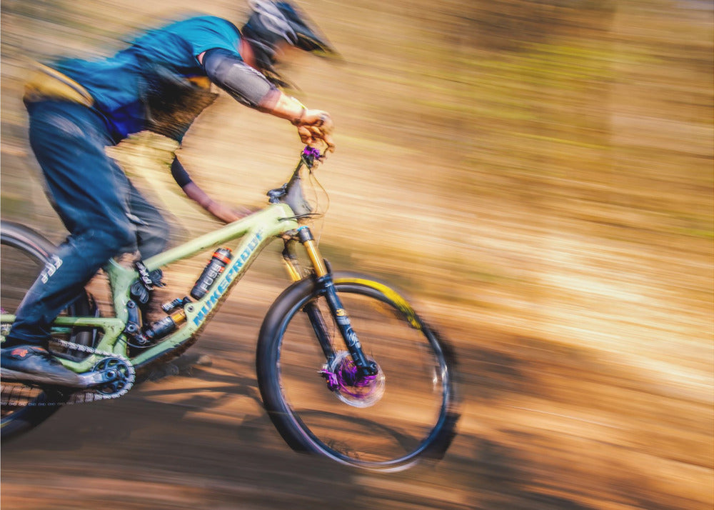 An action shot of a mountain biker in full gear speeding down a dirt trail, captured with a motion blur effect that streaks the brown and green background, conveying a sense of intense speed. Poster