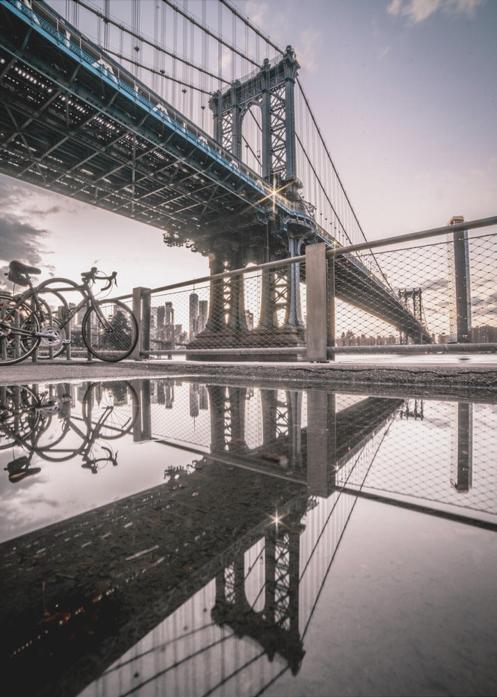 A dramatic low-angle photograph of the Manhattan Bridge, with its intricate steel structure and cables reflected in a large puddle on the pavement in the foreground. Two bicycles are parked near a fence on the left, with the New York City skyline visible in the distance under a softly lit, cloudy sky. Wall Art