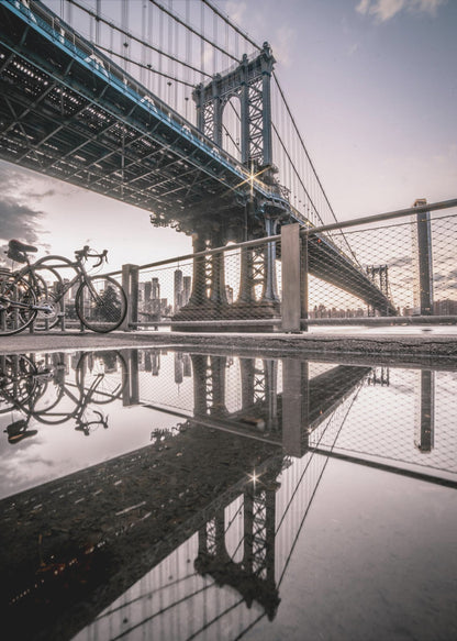 A dramatic low-angle photograph of the Manhattan Bridge, with its intricate steel structure and cables reflected in a large puddle on the pavement in the foreground. Two bicycles are parked near a fence on the left, with the New York City skyline visible in the distance under a softly lit, cloudy sky. Wall Art