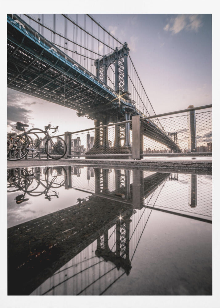 A dramatic low-angle photograph of the Manhattan Bridge, with its intricate steel structure and cables reflected in a large puddle on the pavement in the foreground. Two bicycles are parked near a fence on the left, with the New York City skyline visible in the distance under a softly lit, cloudy sky. Wall Art