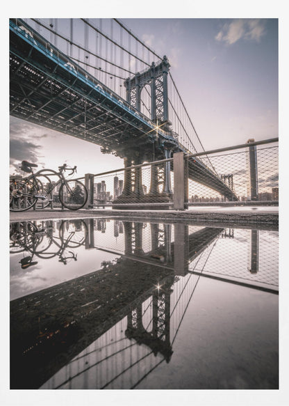 A dramatic low-angle photograph of the Manhattan Bridge, with its intricate steel structure and cables reflected in a large puddle on the pavement in the foreground. Two bicycles are parked near a fence on the left, with the New York City skyline visible in the distance under a softly lit, cloudy sky. Wall Art