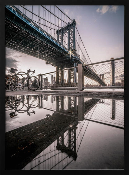 A dramatic low-angle photograph of the Manhattan Bridge, with its intricate steel structure and cables reflected in a large puddle on the pavement in the foreground. Two bicycles are parked near a fence on the left, with the New York City skyline visible in the distance under a softly lit, cloudy sky. Wall Art