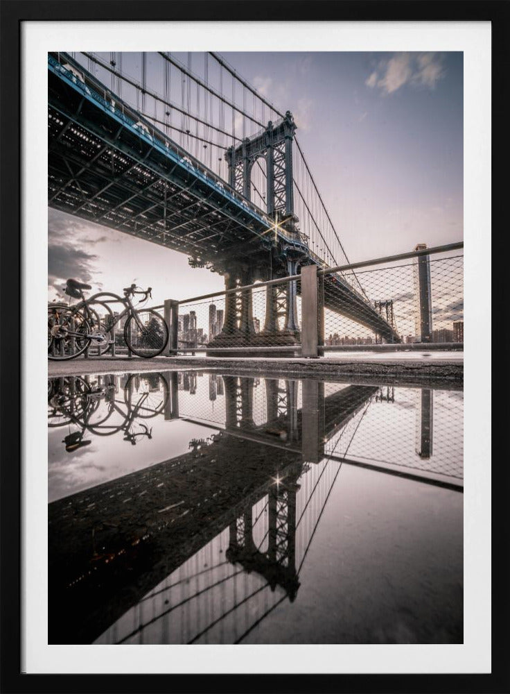 A dramatic low-angle photograph of the Manhattan Bridge, with its intricate steel structure and cables reflected in a large puddle on the pavement in the foreground. Two bicycles are parked near a fence on the left, with the New York City skyline visible in the distance under a softly lit, cloudy sky. Wall Art