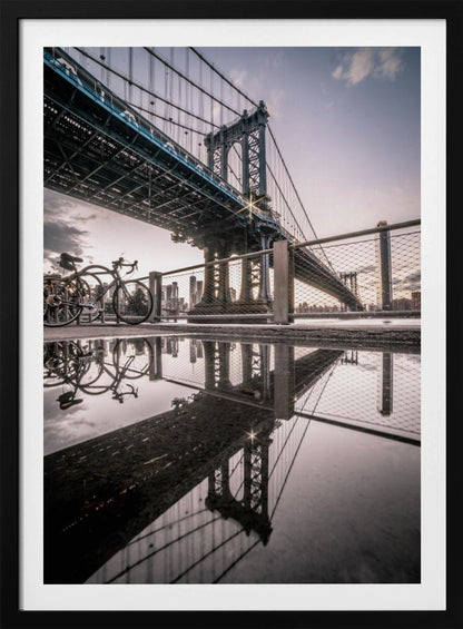 A dramatic low-angle photograph of the Manhattan Bridge, with its intricate steel structure and cables reflected in a large puddle on the pavement in the foreground. Two bicycles are parked near a fence on the left, with the New York City skyline visible in the distance under a softly lit, cloudy sky. Wall Art