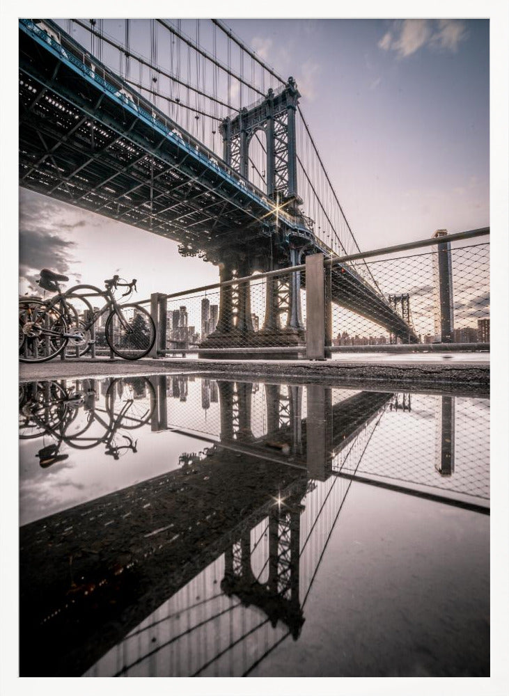A dramatic low-angle photograph of the Manhattan Bridge, with its intricate steel structure and cables reflected in a large puddle on the pavement in the foreground. Two bicycles are parked near a fence on the left, with the New York City skyline visible in the distance under a softly lit, cloudy sky. Wall Art
