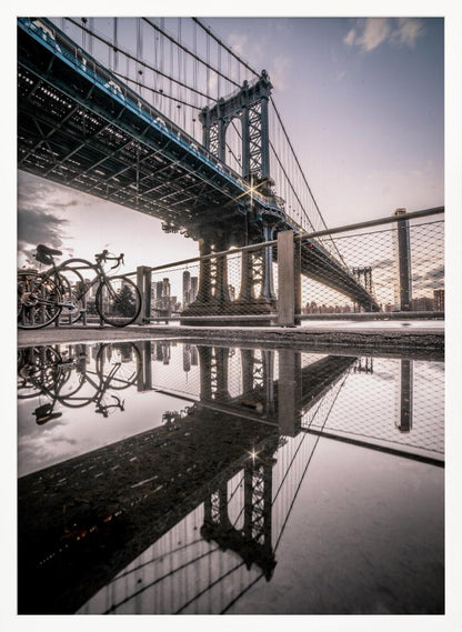 A dramatic low-angle photograph of the Manhattan Bridge, with its intricate steel structure and cables reflected in a large puddle on the pavement in the foreground. Two bicycles are parked near a fence on the left, with the New York City skyline visible in the distance under a softly lit, cloudy sky. Wall Art