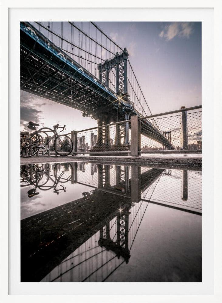A dramatic low-angle photograph of the Manhattan Bridge, with its intricate steel structure and cables reflected in a large puddle on the pavement in the foreground. Two bicycles are parked near a fence on the left, with the New York City skyline visible in the distance under a softly lit, cloudy sky. Wall Art
