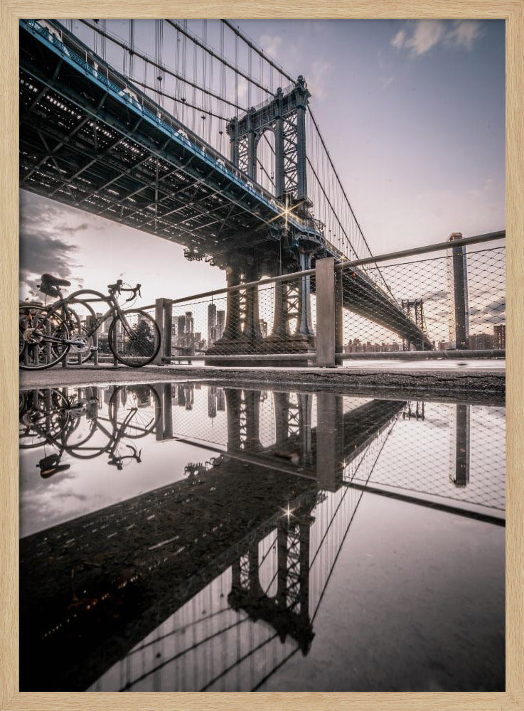 A dramatic low-angle photograph of the Manhattan Bridge, with its intricate steel structure and cables reflected in a large puddle on the pavement in the foreground. Two bicycles are parked near a fence on the left, with the New York City skyline visible in the distance under a softly lit, cloudy sky. Wall Art