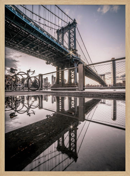 A dramatic low-angle photograph of the Manhattan Bridge, with its intricate steel structure and cables reflected in a large puddle on the pavement in the foreground. Two bicycles are parked near a fence on the left, with the New York City skyline visible in the distance under a softly lit, cloudy sky. Wall Art