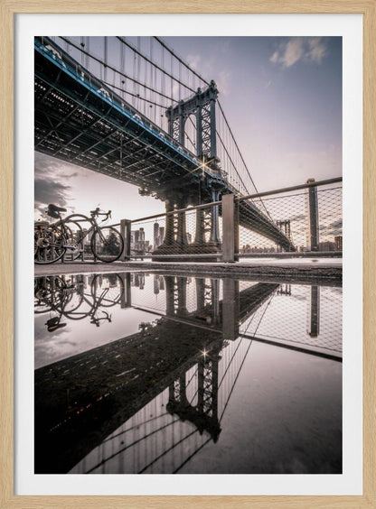A dramatic low-angle photograph of the Manhattan Bridge, with its intricate steel structure and cables reflected in a large puddle on the pavement in the foreground. Two bicycles are parked near a fence on the left, with the New York City skyline visible in the distance under a softly lit, cloudy sky. Wall Art