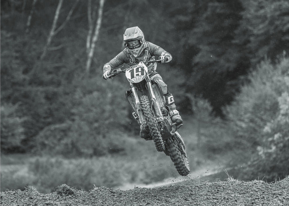 A dynamic black and white photograph of a motocross racer in mid-air, jumping over a dirt mound. The rider, wearing full protective gear and a helmet, is on a dirt bike with the number 19 plate. The background is a soft-focus forest, and the entire image is enclosed in a silver frame. Poster