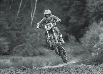 A dynamic black and white photograph of a motocross racer in mid-air, jumping over a dirt mound. The rider, wearing full protective gear and a helmet, is on a dirt bike with the number 19 plate. The background is a soft-focus forest, and the entire image is enclosed in a silver frame. Poster