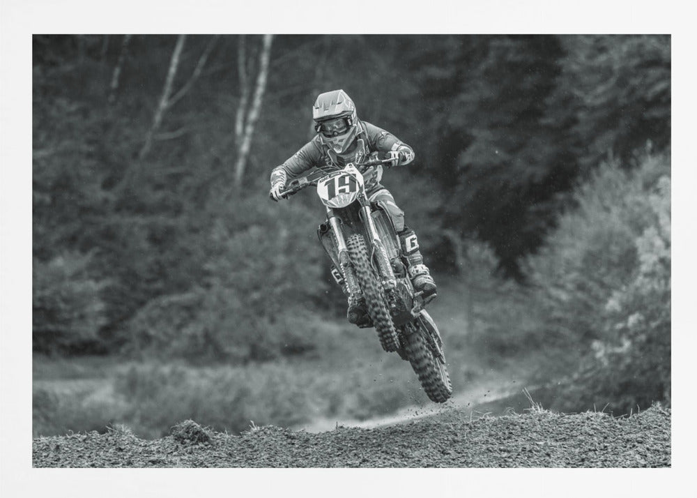 A dynamic black and white photograph of a motocross racer in mid-air, jumping over a dirt mound. The rider, wearing full protective gear and a helmet, is on a dirt bike with the number 19 plate. The background is a soft-focus forest, and the entire image is enclosed in a silver frame. Poster