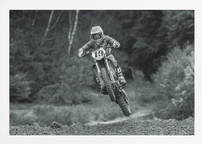 A dynamic black and white photograph of a motocross racer in mid-air, jumping over a dirt mound. The rider, wearing full protective gear and a helmet, is on a dirt bike with the number 19 plate. The background is a soft-focus forest, and the entire image is enclosed in a silver frame. Poster